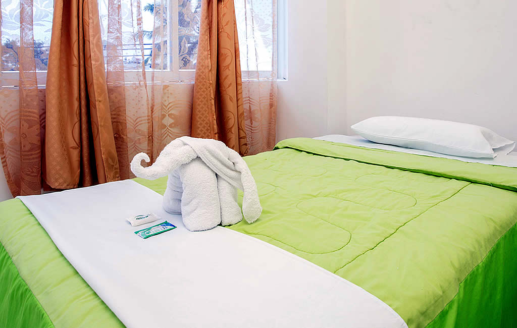 A neatly made bed dressed in a green and white coverlet features a decorative towel folded into an elephant shape. Natural light filters through sheer curtains, illuminating the room, complemented by soft orange drapes that frame the window.