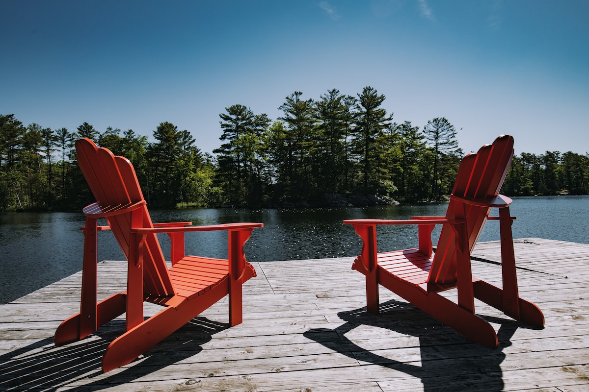 Two bright red Adirondack chairs are positioned on a wooden dock, facing a serene lake. The surrounding landscape includes a backdrop of lush green trees under a clear blue sky, creating a peaceful atmosphere.