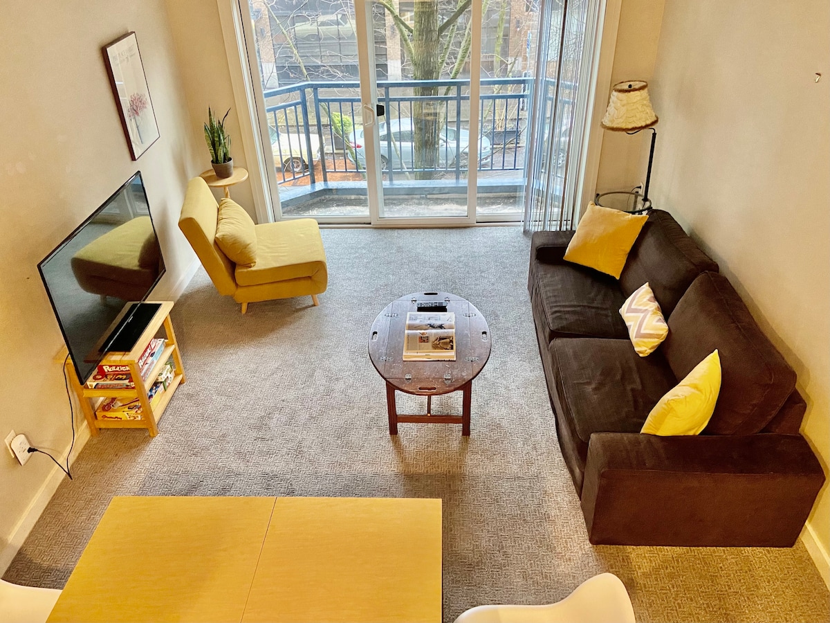A cozy living area is presented, featuring a brown sofa with yellow accent pillows and a yellow chair beside a round wooden coffee table. A television is positioned on a small stand, and natural light enters through a large balcony door, enhancing the relaxed atmosphere.