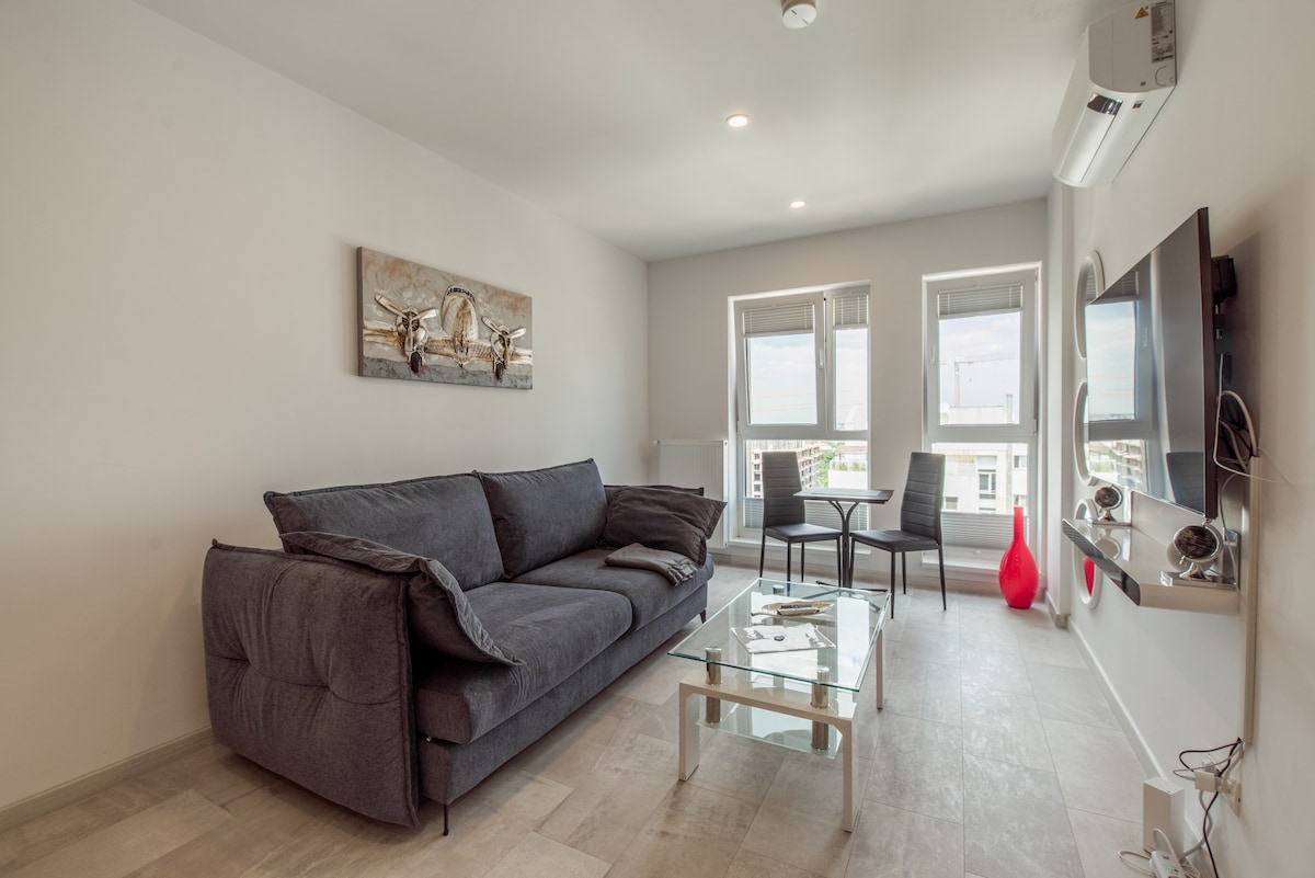 A modern living area features a gray fabric sofa and a glass coffee table. Two black chairs are positioned around the table, while a minimalistic wall art piece is displayed above the sofa. Natural light enters through the large windows.