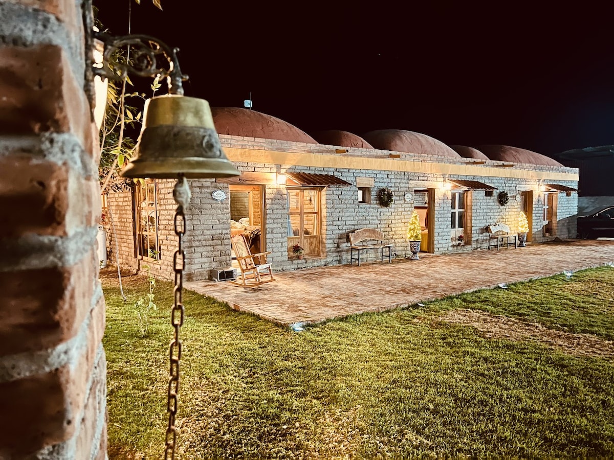 A rustic property with a stone exterior features curved rooftops, illuminated gently by soft outdoor lighting. A wooden rocking chair and benches are placed on a grassy area in front, inviting relaxation. The peaceful evening atmosphere highlights the charm of the retreat.
