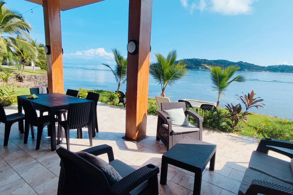 A shaded outdoor terrace features a spacious dining table surrounded by black chairs. Comfortable seating areas are arranged nearby, offering views of the clear blue water and lush greenery, enhancing the relaxing atmosphere of the space.