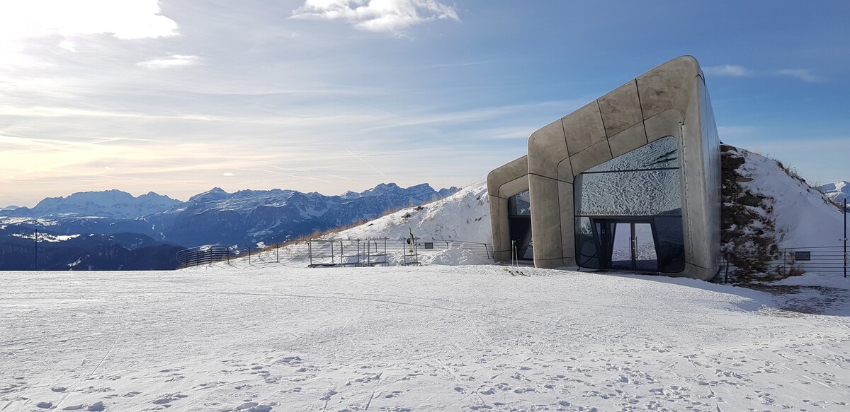 A modern architectural structure sits on snow-covered terrain, surrounded by majestic mountain vistas. The building features large glass panels and a unique angular design, contrasting with the serene winter landscape under a clear blue sky.