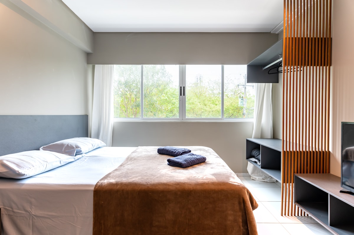 A spacious sleeping area features a large bed with neutral bedding and two towels neatly arranged on top. The room is illuminated by natural light streaming through large windows adorned with sheer curtains. A wooden accent wall and shelving unit add a modern touch.