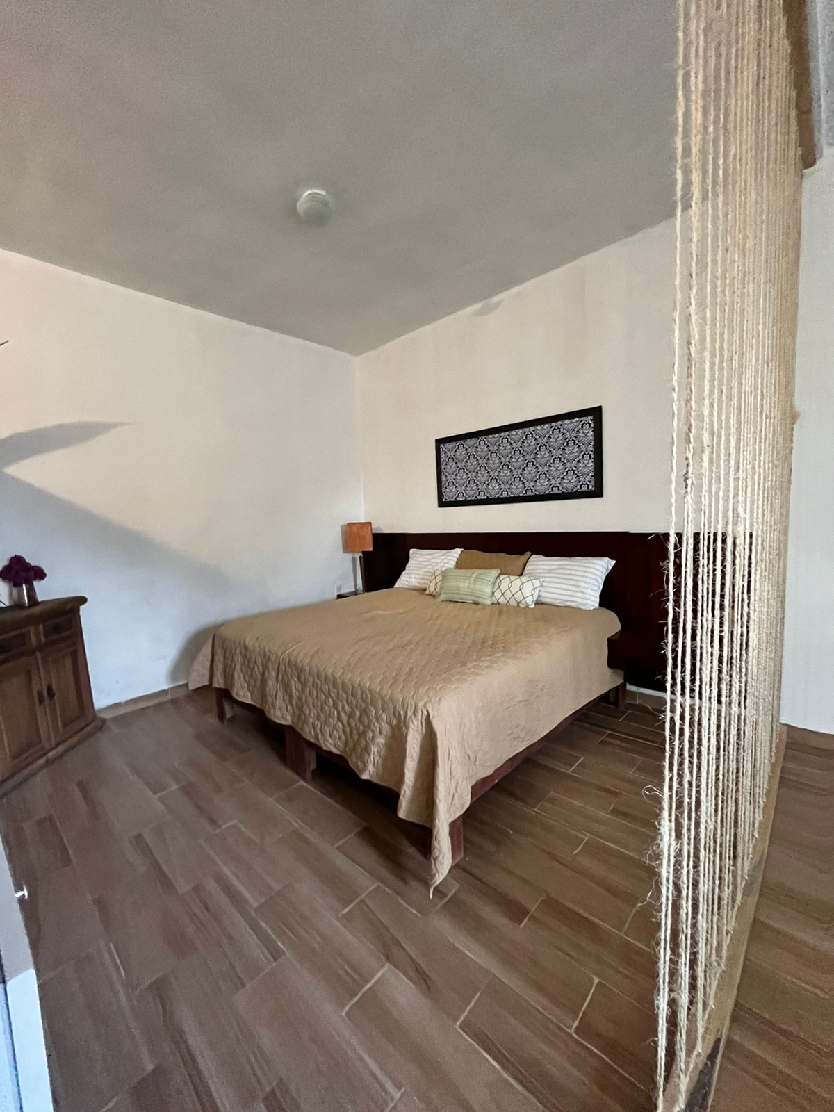 A spacious bedroom features a large bed with neutral bedding and accent pillows. Natural light enters through a window, which is framed by a simple decorative curtain. A wooden dresser is positioned against one wall, while a decorative piece hangs above the bed.