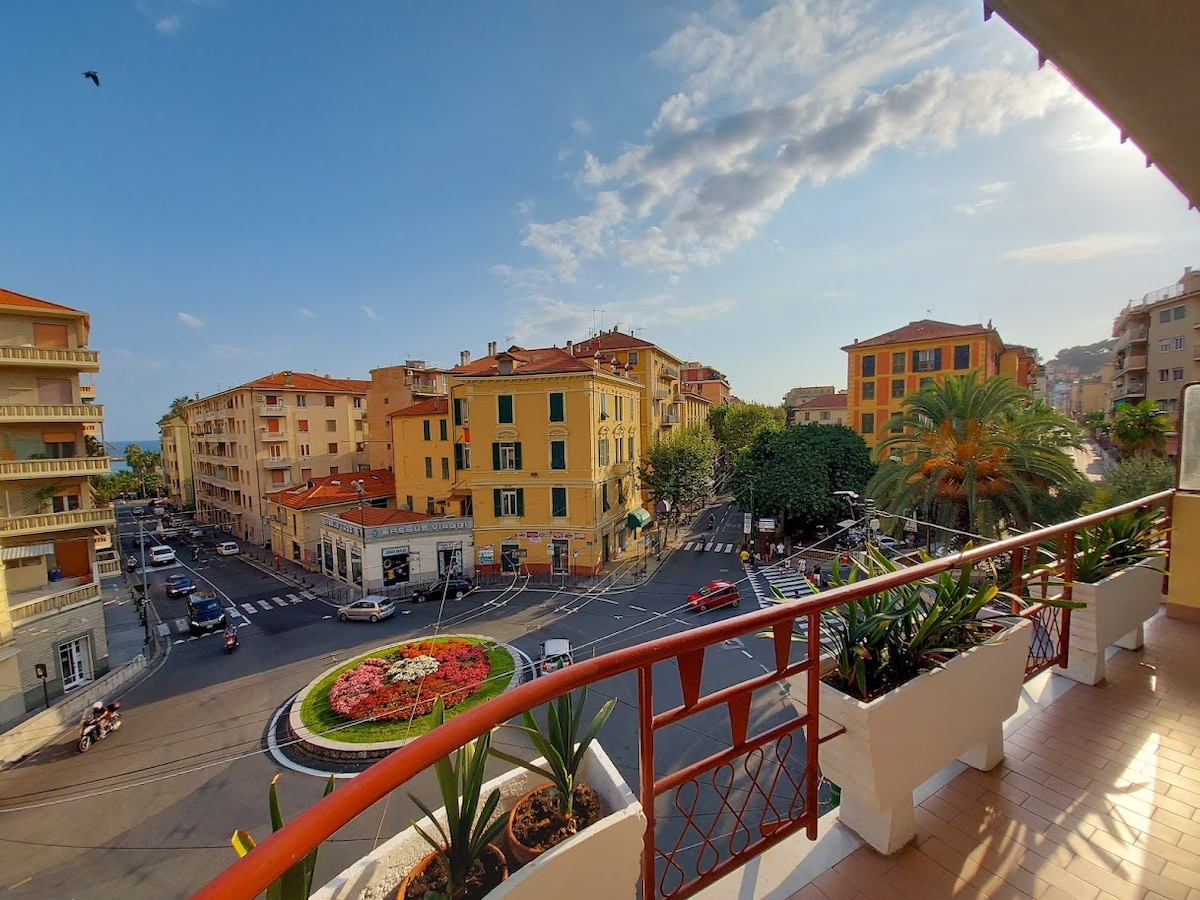 A view from the balcony showcases a lively street intersection adorned with a circular flowerbed. Nearby buildings exhibit warm colors, while palm trees add greenery to the scene. The horizon features the sky with occasional clouds, hinting at a pleasant day.