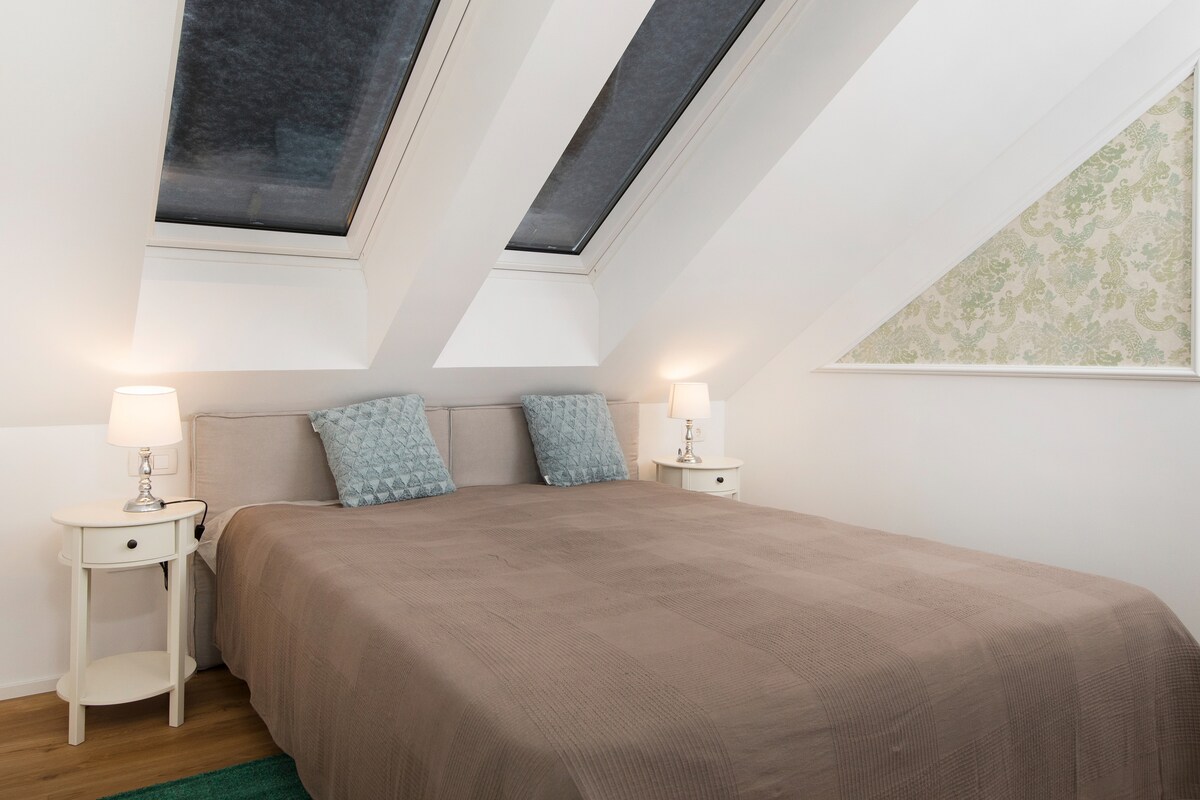 A cozy bedroom features a spacious bed dressed in neutral linens, flanked by two bedside tables with lamps. Natural light enters through skylights, illuminating the minimalist decor, including a patterned wall in soft colors.