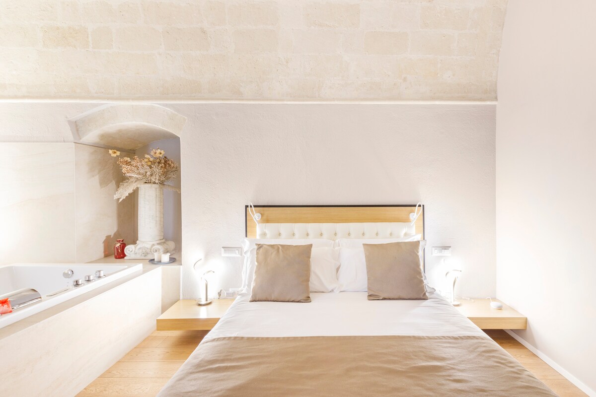 A spacious bed is positioned against a softly lit wall with two bedside lamps, providing ample light for reading. A plush throw is draped over the bed, and a modern bathroom area is visible in the background, featuring a relaxing bathtub and decorative accents.