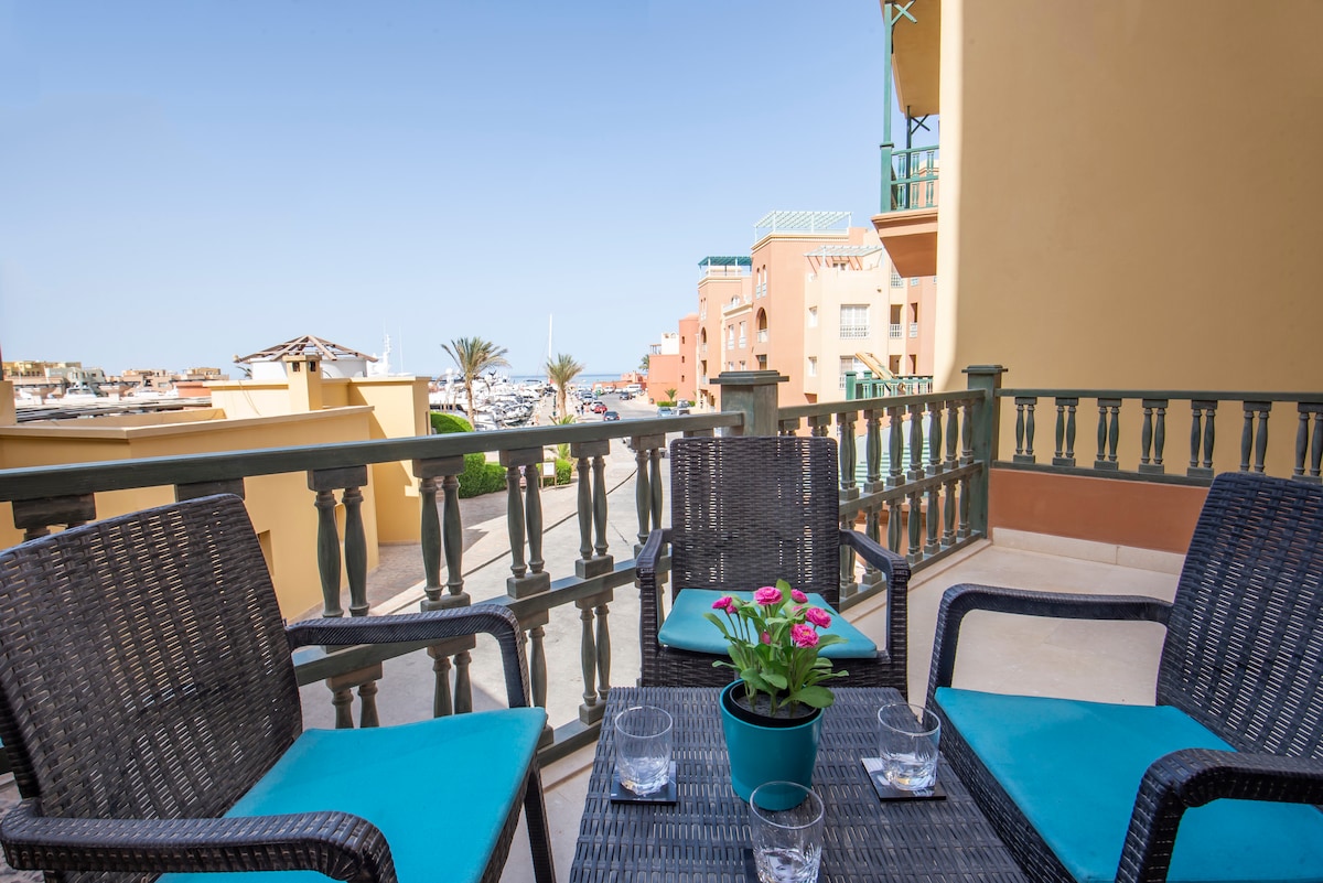 A balcony seating area is furnished with three wicker chairs, each adorned with bright blue cushions. A small potted plant sits on a table between the chairs, creating a relaxed outdoor space overlooking the marina and lined with palm trees.