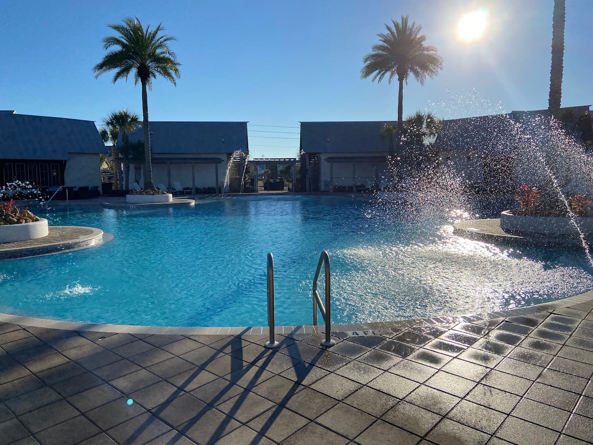A large pool is seen surrounded by palm trees, with sunlight reflecting on the water's surface. Water features create gentle splashes at the pool's edge, while spacious areas for lounging are visible around the pool deck.