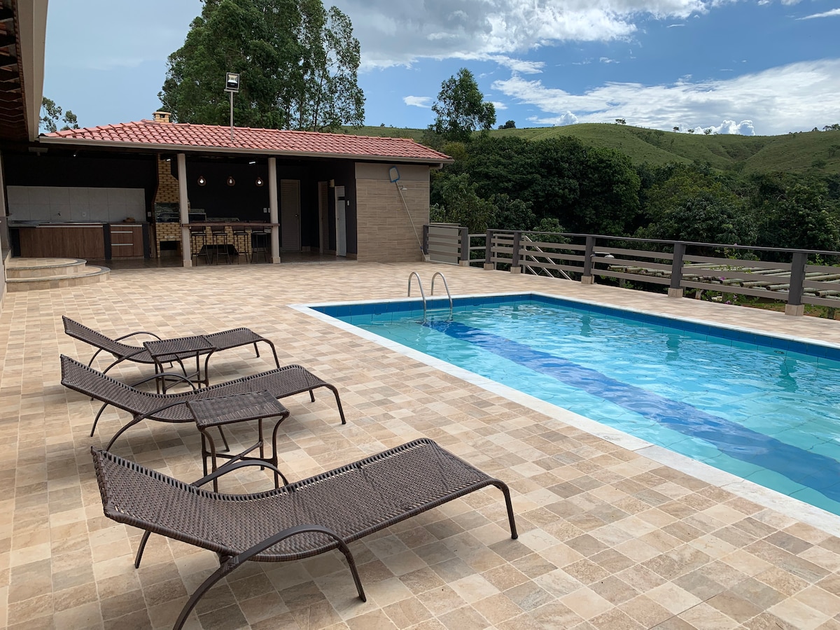 A spacious outdoor area features a rectangular swimming pool surrounded by a tiled deck. Four lounge chairs are positioned nearby, offering a place to relax. A covered grilling area with seating is visible in the background, framed by lush greenery and distant hills.