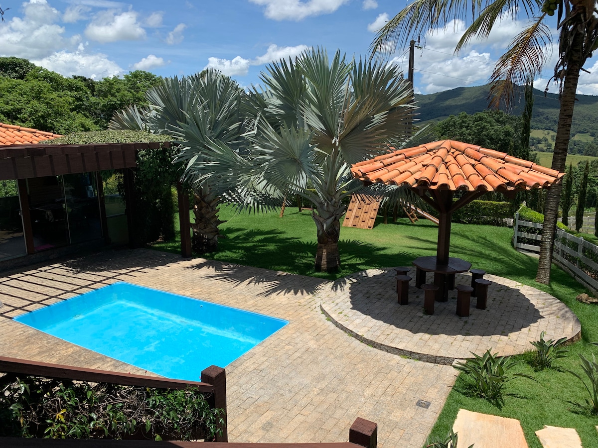 A tranquil outdoor area features a blue swimming pool surrounded by landscaped gardens. A covered seating area with a tiled roof and wooden tables is positioned nearby, with palm trees providing shade. The scenic backdrop includes rolling hills under a clear sky.