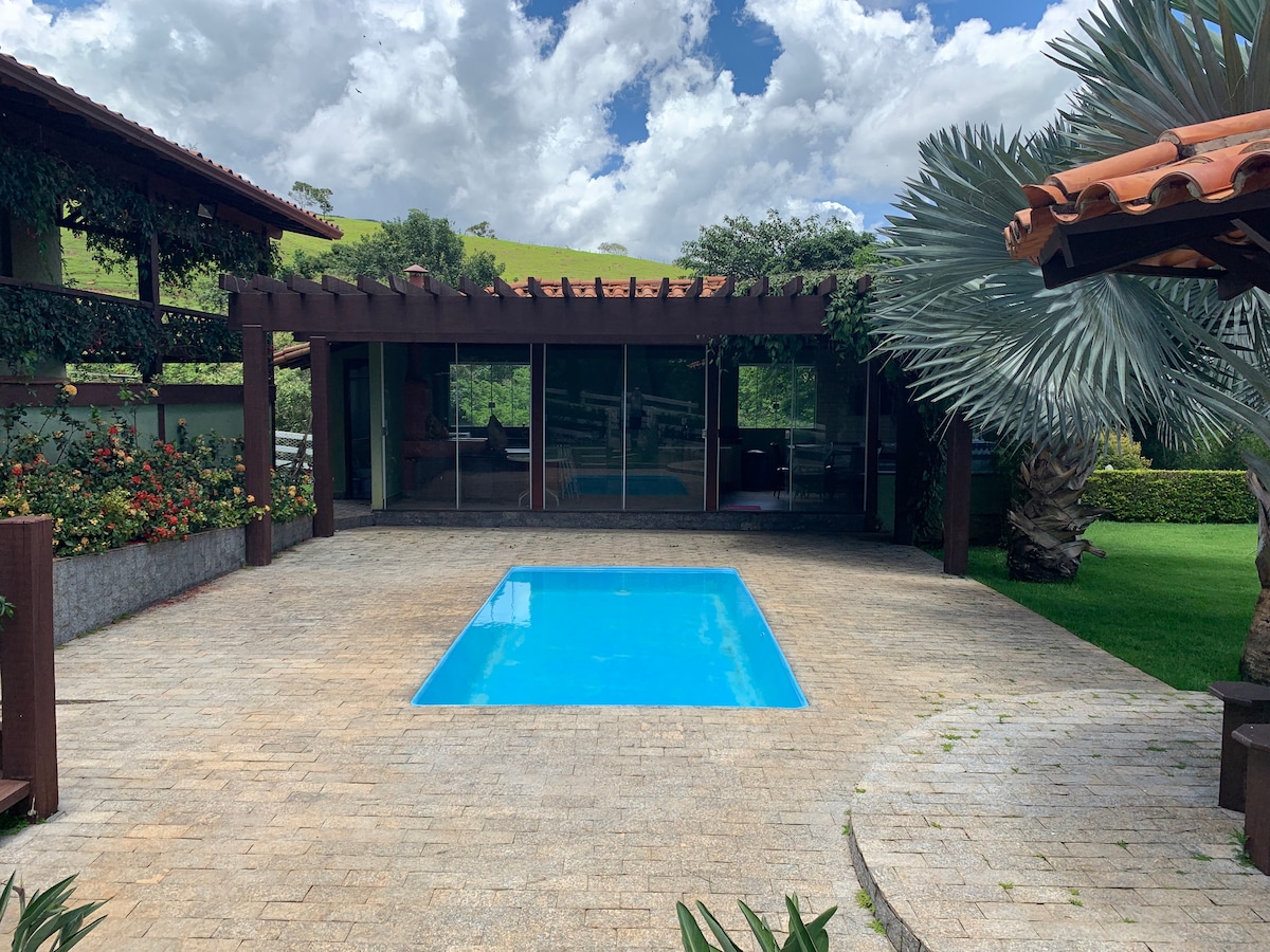 A clear blue pool is situated within a stone patio, surrounded by well-maintained greenery and flowers. The modern structure features large glass walls, providing views of the tranquil outdoor landscape. A pergola provides shade, enhancing the outdoor space for relaxation.