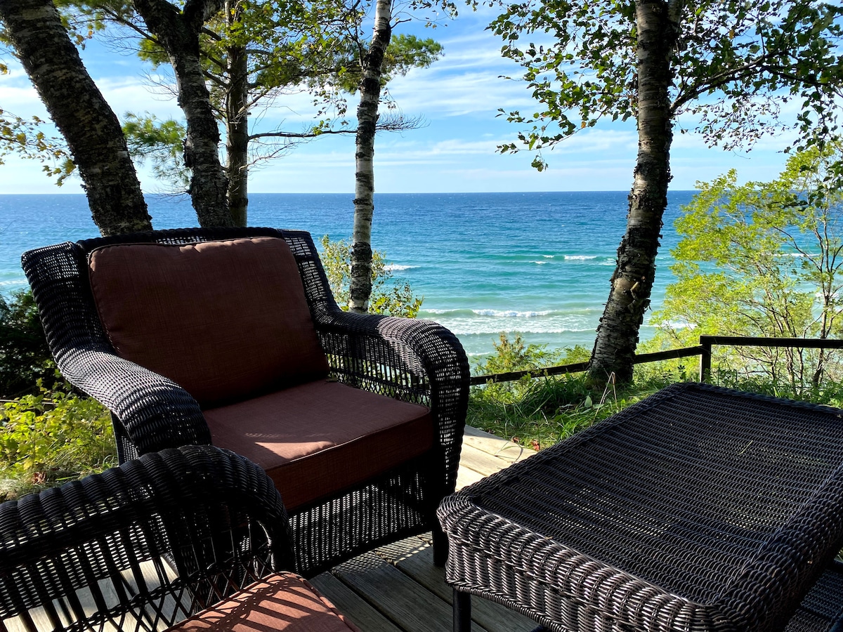 Seating is provided on a deck overlooking Lake Michigan, with two wicker chairs and a small table. The tranquil blue waters and gentle waves can be seen through the trees, creating a serene coastal setting.