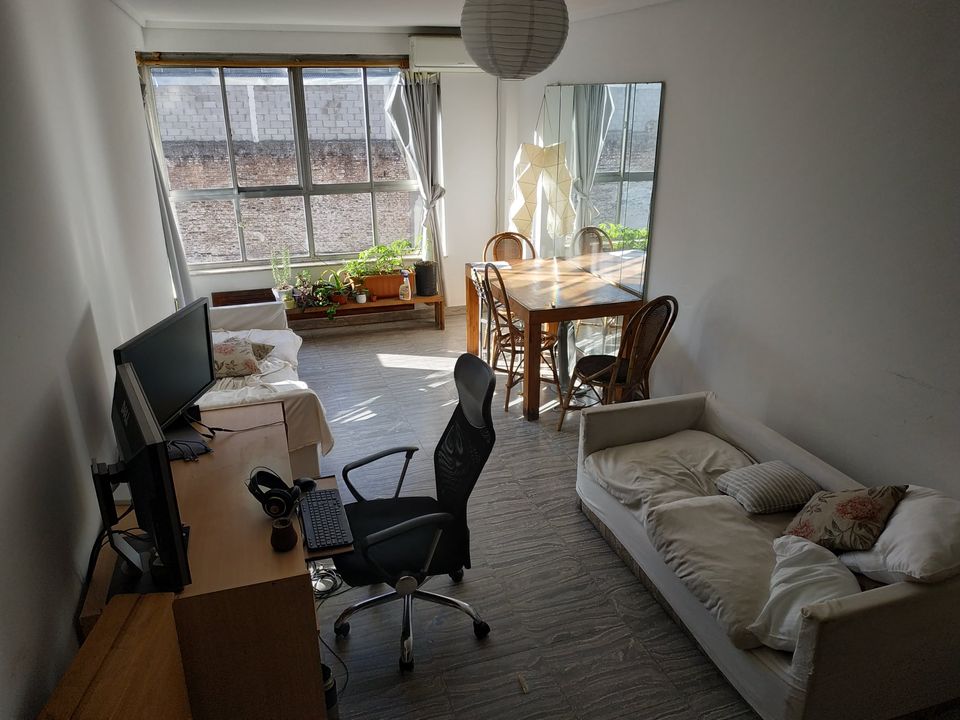 A well-lit living area features large windows allowing natural light to flood the space. A dining table with wooden chairs is positioned near the window. A comfortable desk with an office chair and a television are also visible, alongside a cozy seating area with a soft daybed.