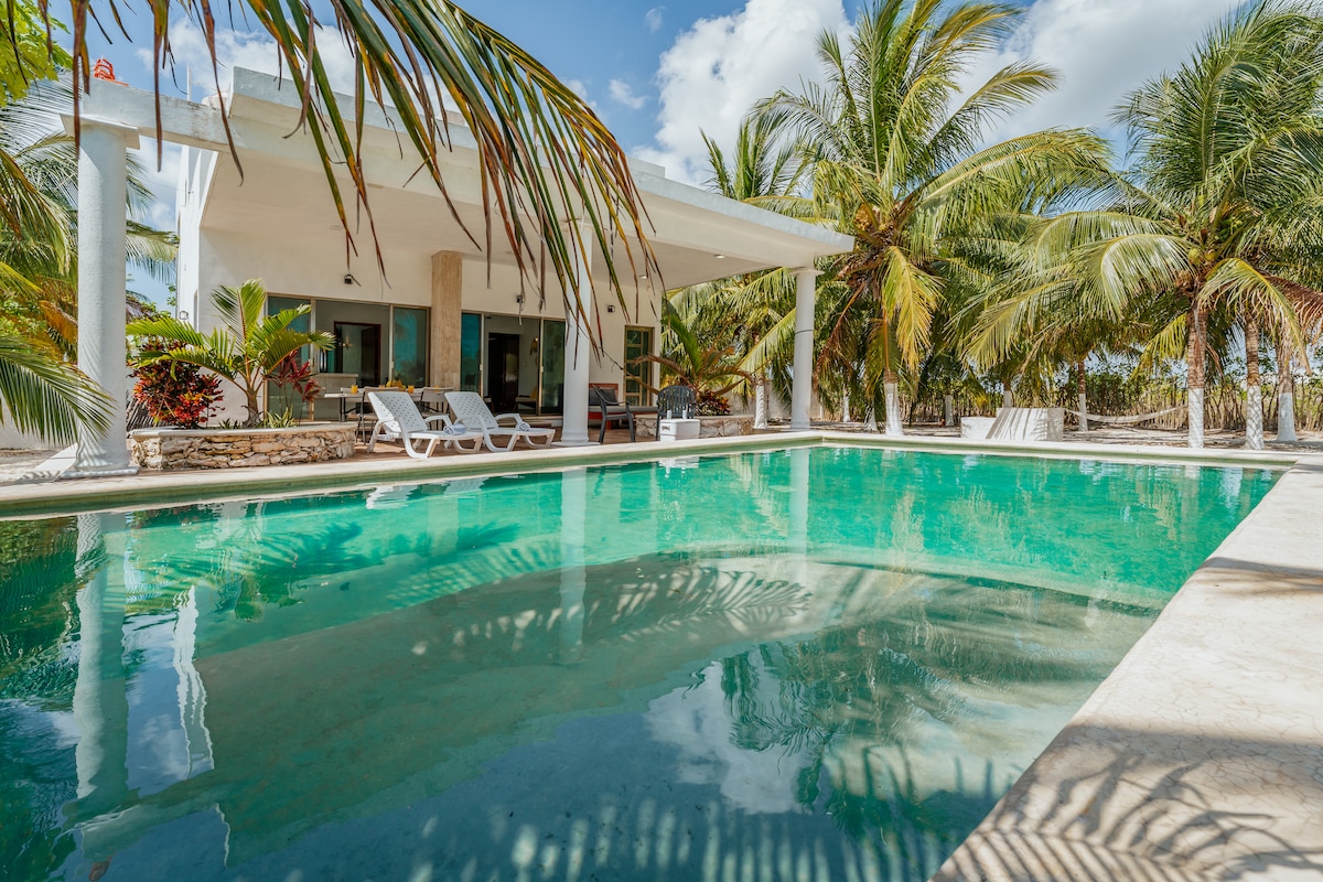 A spacious swimming pool reflects the surrounding palm trees and sky. Lounge chairs are positioned by the poolside, inviting relaxation. The covered terrace with a light-colored overhang offers shade, creating a serene environment for enjoyment and leisure.