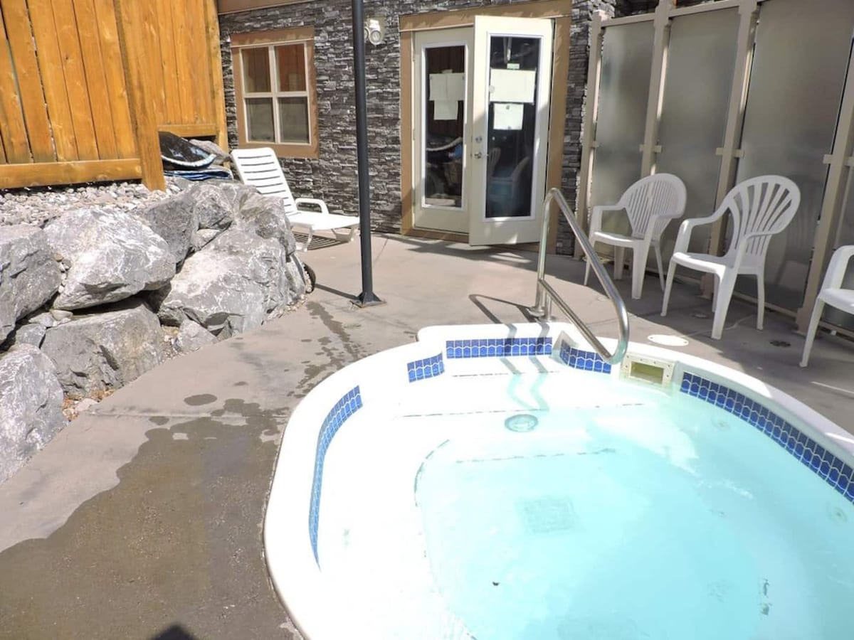 A private hot tub is set on a stone patio, surrounded by large rocks and wooden fencing. Light glistens on the water surface, while two white chairs are positioned nearby. Glass doors lead to an indoor space, partially obscured by frosted glass panels.