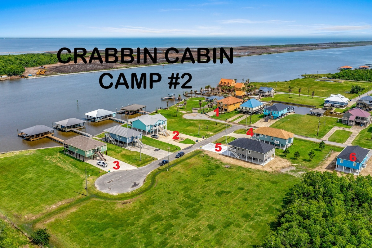 An aerial view displays a waterfront community with colorful homes and gathering spots. The adjacent pier and waterway are visible, with expansive green lawns providing space for outdoor activities. Notable structures include shaded boat lifts and open spaces, facilitating access to crabbing and fishing.