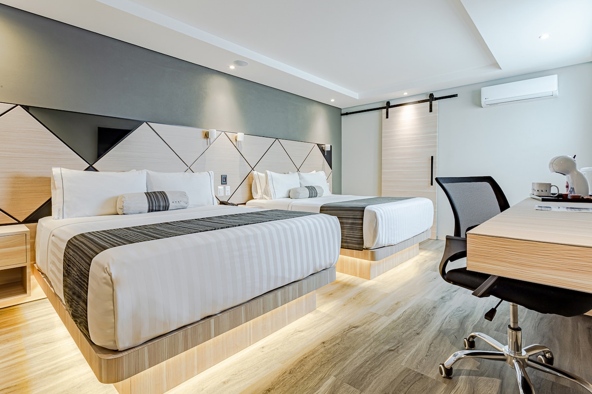 The room features two spacious beds with light bedding, positioned by a decorative headboard. A modern workspace with a chair is situated near a window. Soft lighting highlights the room's contemporary design and wood flooring, while an air conditioning unit is mounted on one wall.