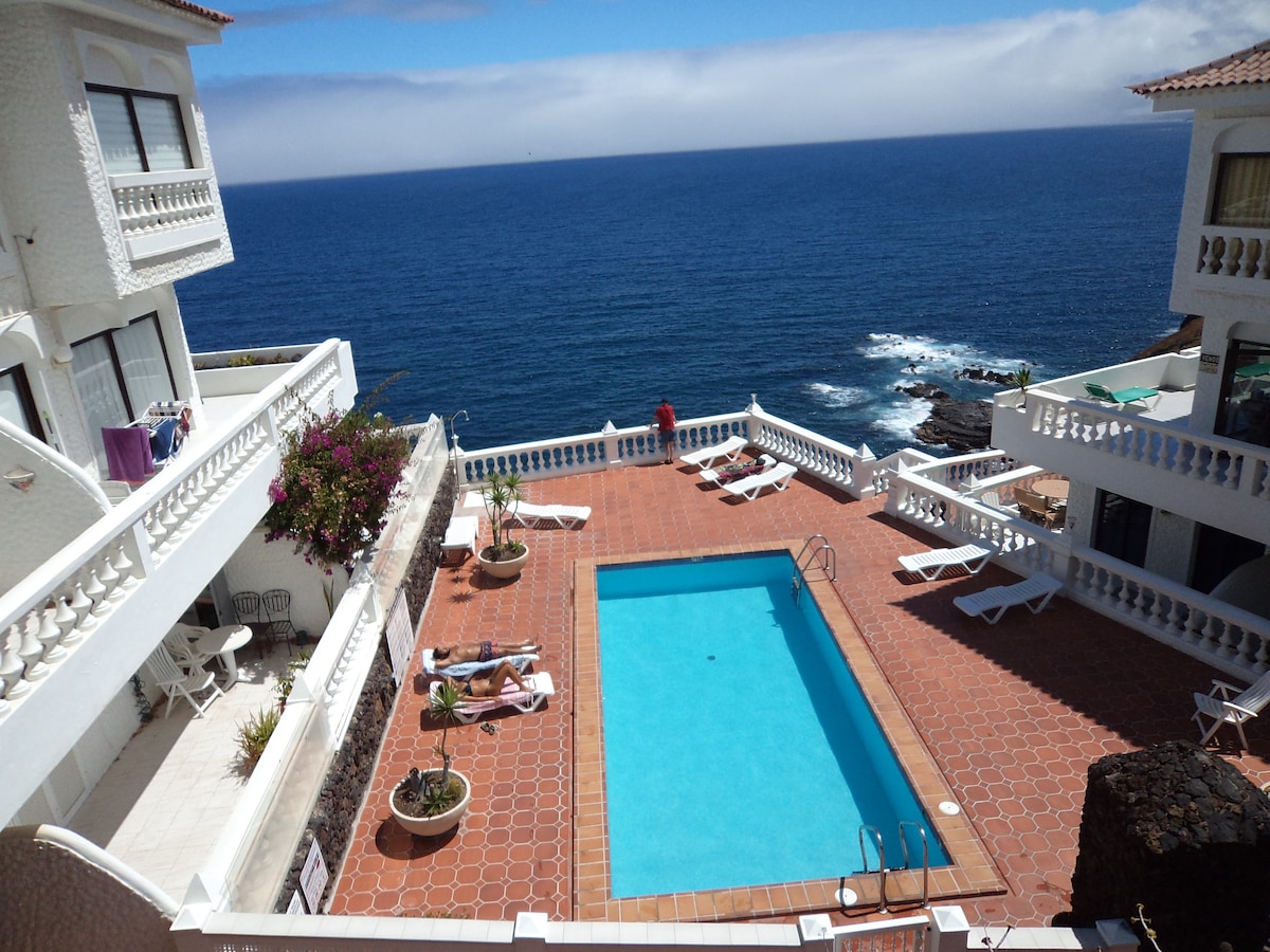 A sunny pool area is viewed from above, featuring a rectangular swimming pool surrounded by tiled flooring. Several lounge chairs are positioned nearby, and colorful potted plants add greenery. The blue sea stretches in the background, with rocky coastline visible.