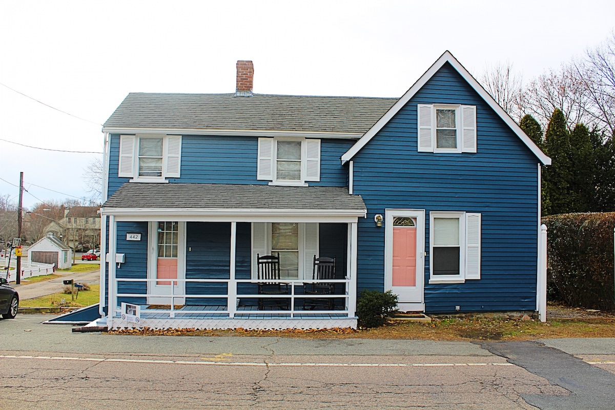 A blue-painted two-story house is shown from the front, featuring a welcoming porch with two rocking chairs. The façade displays white shutters and doors, with one door painted pink. An open area is visible in front of the house, accompanied by a landscaped border.