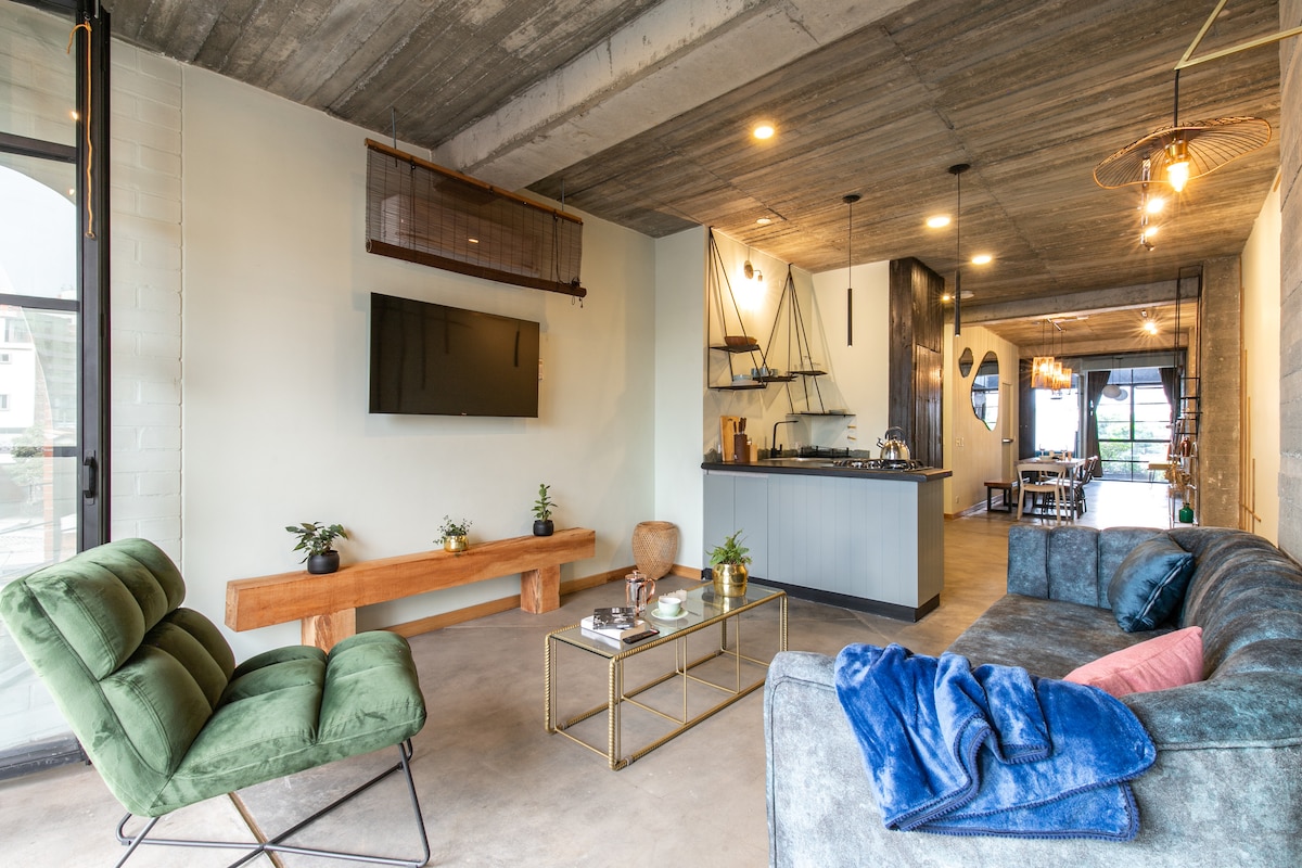 A spacious living area features a large sofa and a green armchair, complemented by a wooden coffee table and a wall-mounted television. The ceiling showcases industrial-style lighting, and large windows allow natural light to fill the space, connecting it to the kitchen and dining area.