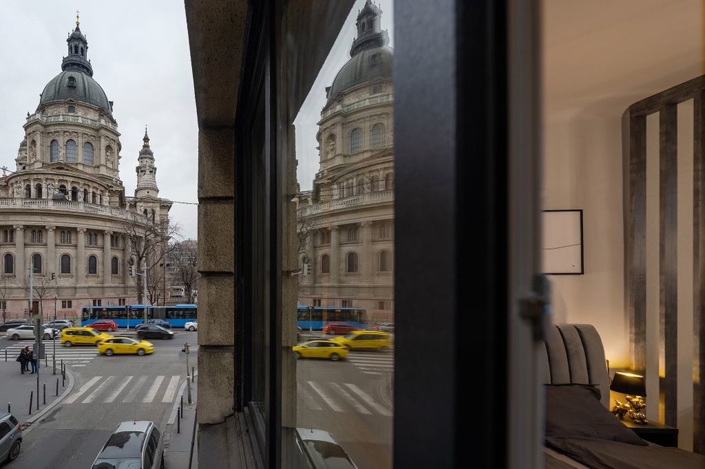 The reflection of the Szent István Basilica is visible in the window, showcasing its grand architecture. Below, yellow taxis pass by on the street, while a cozy sleeping area is seen through the window, featuring neutral-colored bedding.