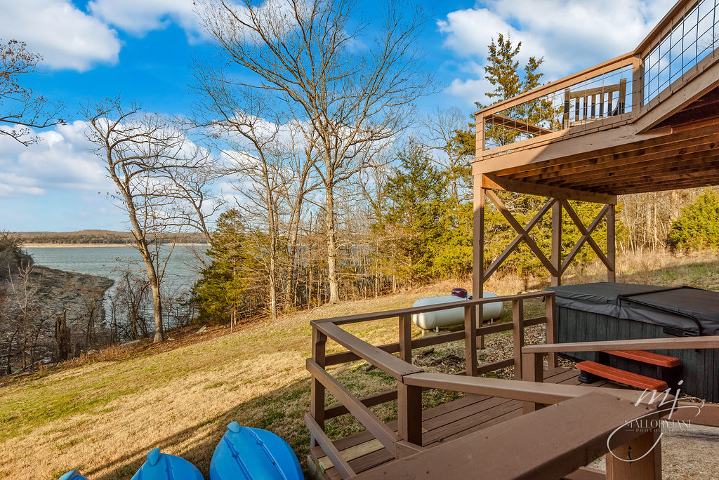 A scenic view of the lake is framed by trees from the spacious outdoor deck. A hot tub is positioned nearby, with blue kayaks resting beside it. The gentle slope of the yard leads down toward the water's edge, enhancing the serene lakeside atmosphere.