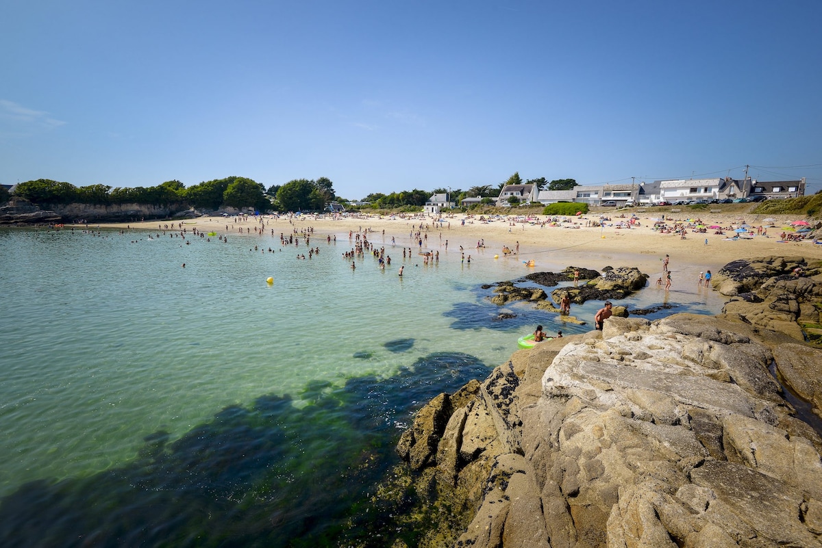 A sandy beach is depicted with gentle waves lapping at the shore. Groups of people are seen enjoying the sun and water, while the rocky outcrop leads into clear waters. Coastal buildings are visible in the background, surrounded by lush greenery.