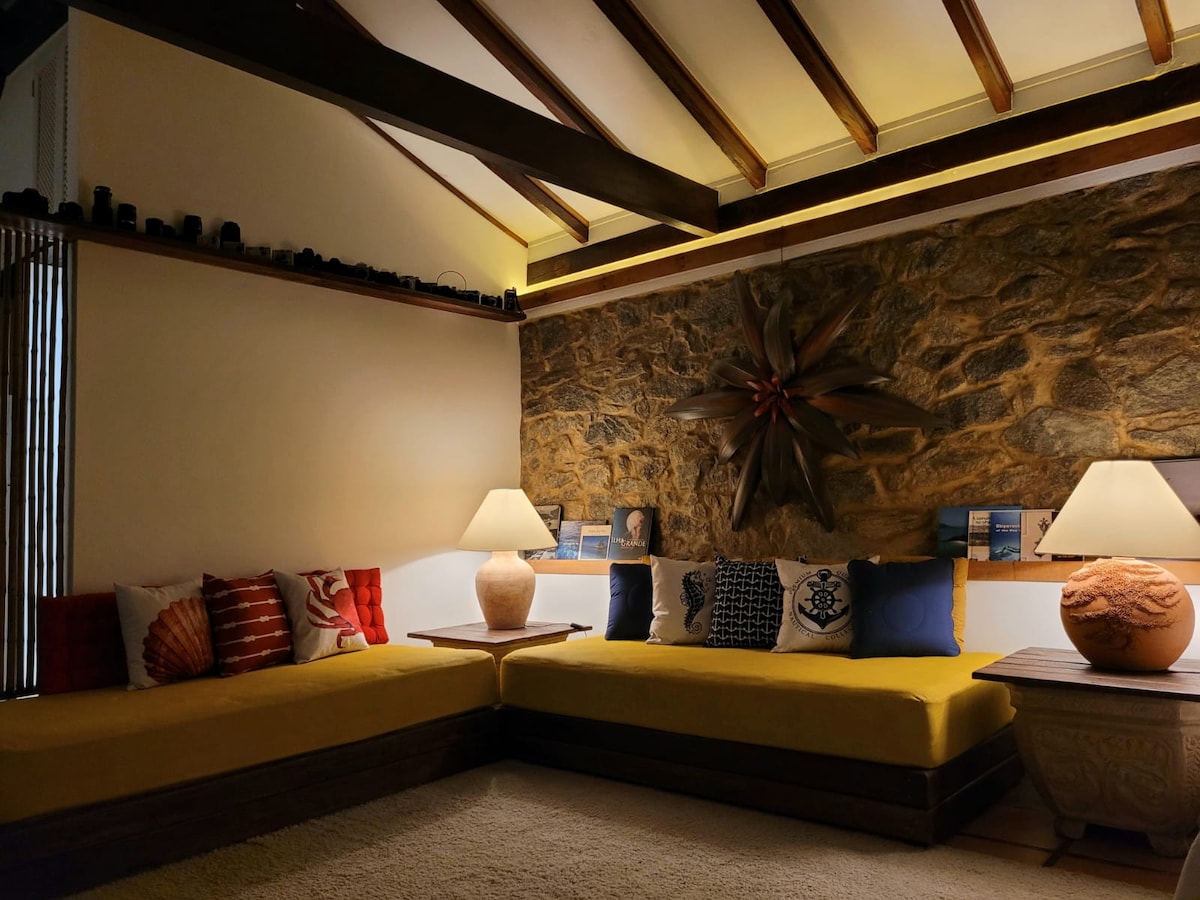 A cozy seating area is featured with a yellow upholstered couch and decorative pillows in various colors. Soft lighting highlights a textured stone wall, while two lamps provide warm illumination. Books are neatly displayed on a ledge, adding to the inviting ambiance.