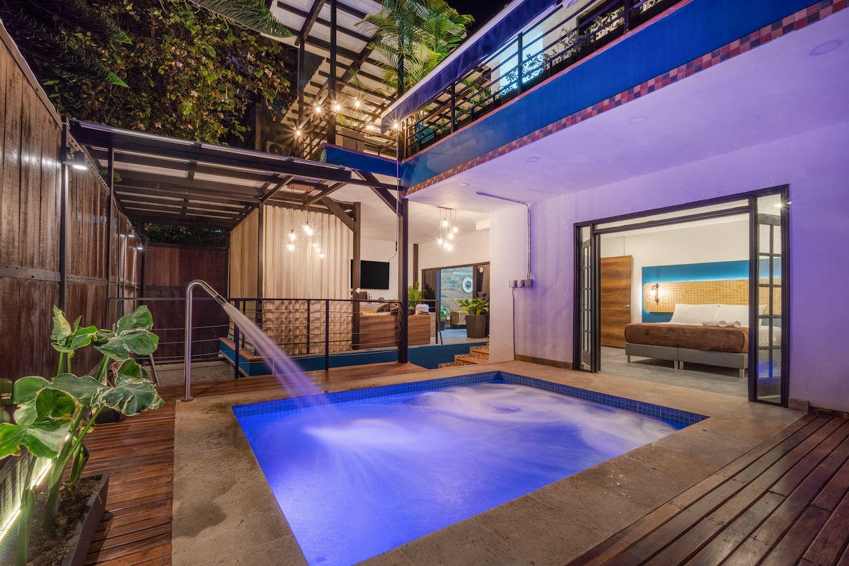 A private outdoor area is highlighted by a spacious Jacuzzi with jets, surrounded by wooden decking. Soft lighting enhances the ambiance, while lush greenery adds a natural element. Nearby, the open indoor living space is visible, connecting seamlessly to the outdoor zone.