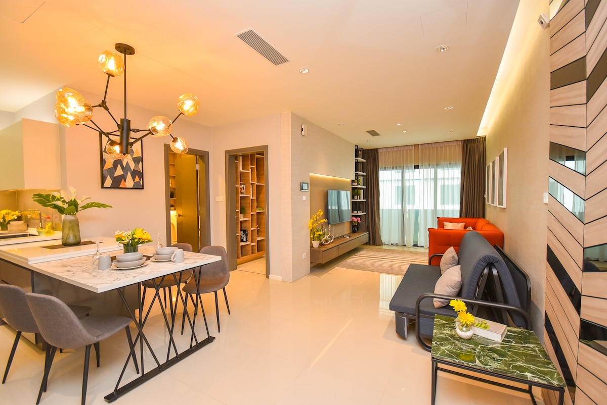 A modern living area features a stylish seating arrangement with a gray sofa and an orange armchair. A dining table is set for four, complemented by decorative flowers. Floor-to-ceiling windows allow natural light to fill the space, while a mounted flat-screen television adds to the contemporary design.