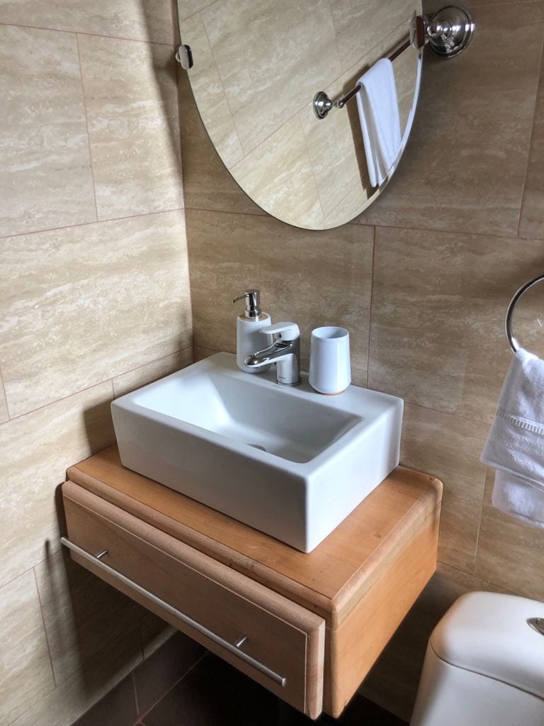 A modern bathroom sink is mounted on a wooden vanity, featuring a clean, rectangular basin. A round mirror reflects the space, while a soap dispenser and a cup are neatly placed beside the sink. White towels hang on a chrome rack nearby.