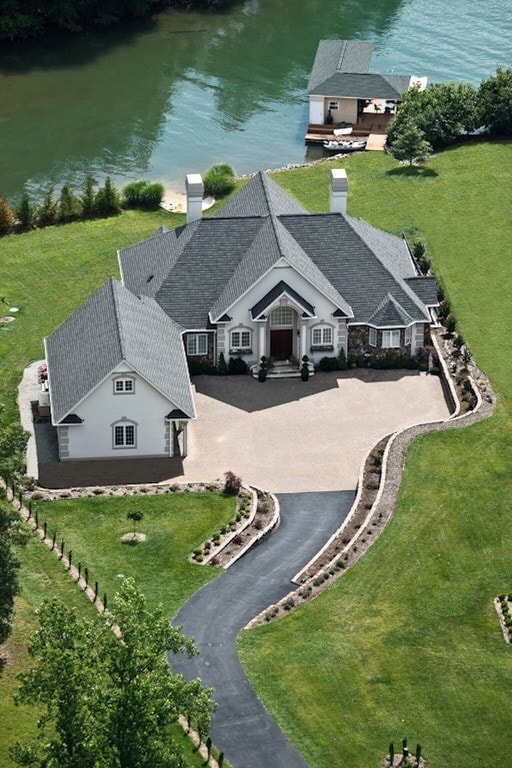 An aerial view showcases a charming two-level home with a manicured lawn and a winding driveway leading to the entrance. The property is situated near a serene body of water, with a dock and additional structure visible in the background, surrounded by lush greenery.