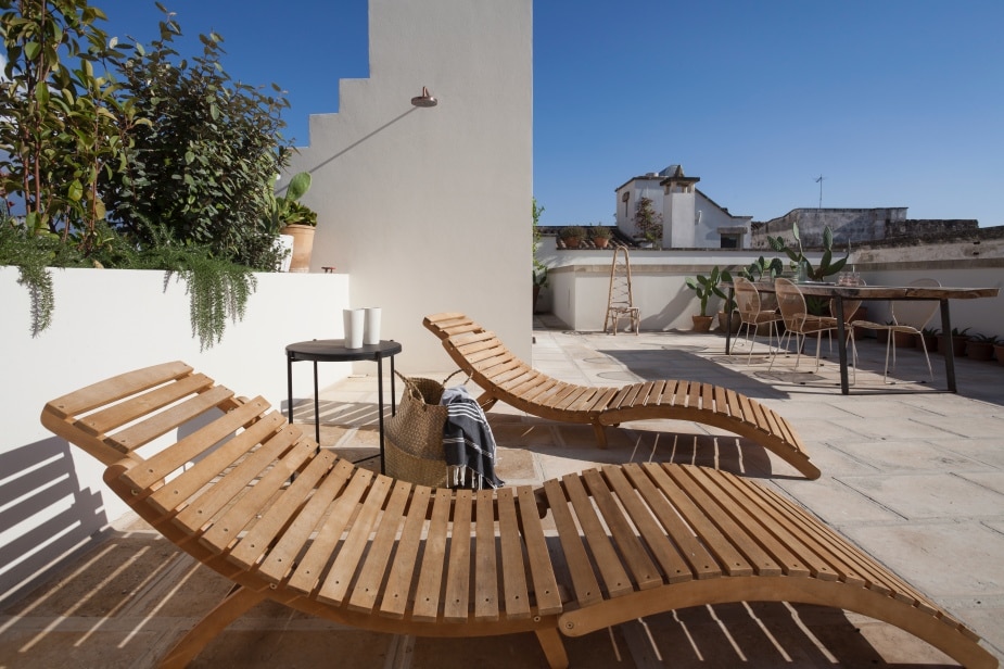 Two wooden lounge chairs are positioned on a spacious terrace, surrounded by potted plants. A small side table sits between them, complementing a soft throw draped over one chair. The terrace offers clear blue skies and views of the surrounding architecture.