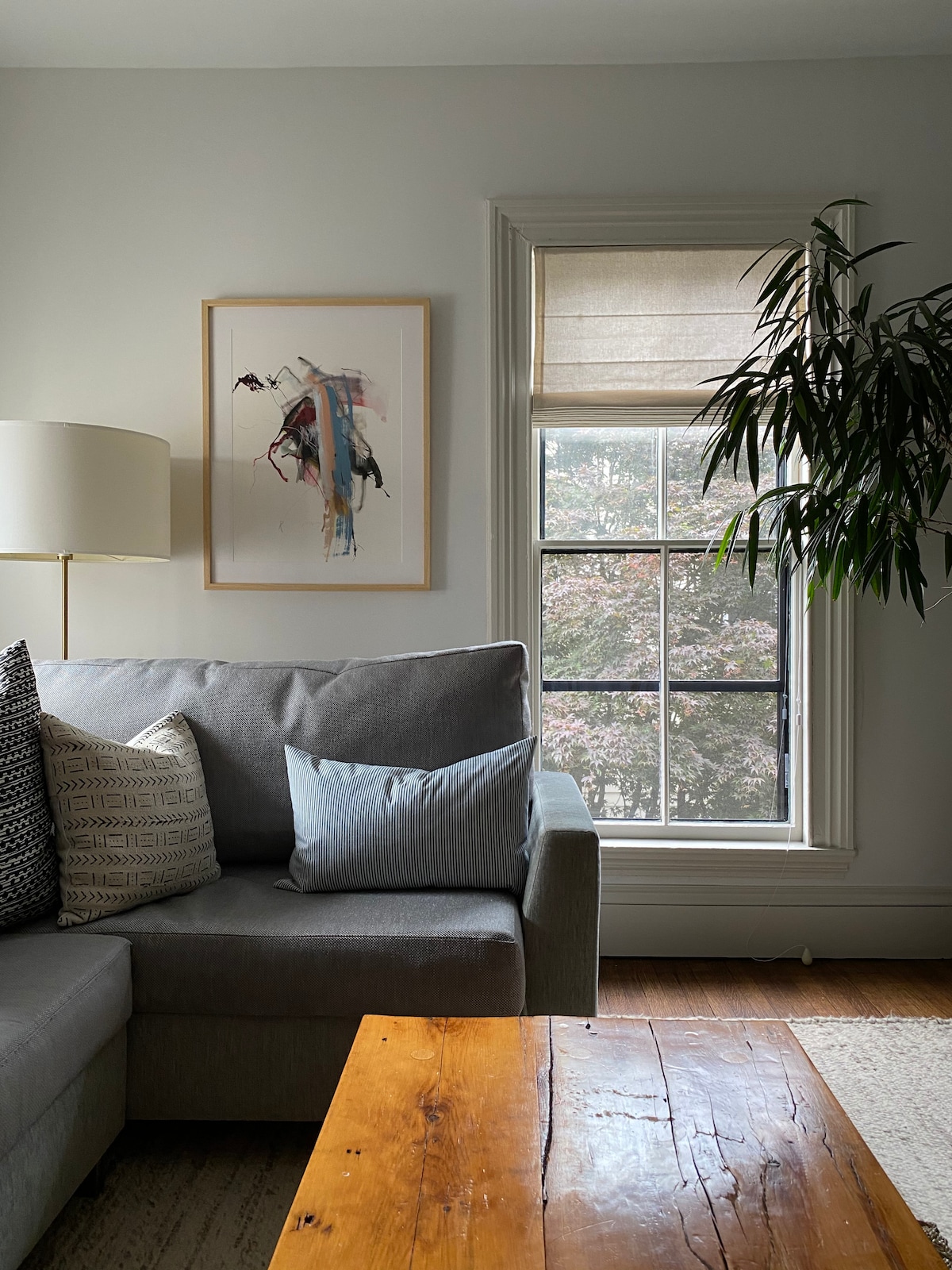 A cozy living area features a grey sectional sofa adorned with decorative pillows. A wooden coffee table sits in front, and a large window allows natural light to fill the space, offering a view of the trees outside. A piece of modern art hangs on the wall.
