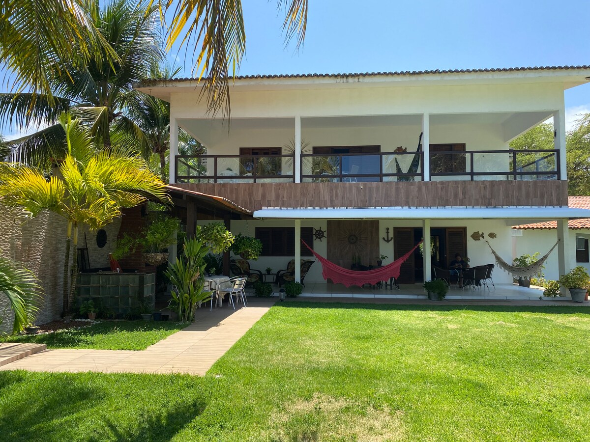 The two-story house features a spacious balcony adorned with a hammock, surrounded by tropical plants. The well-maintained yard showcases lush green grass and a pathway leading to outdoor seating. Large windows provide ample natural light, enhancing the welcoming appearance of the property.
