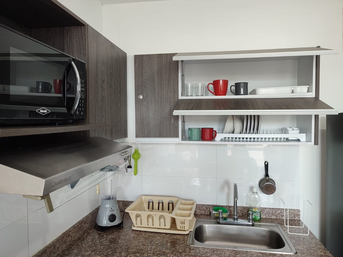 The kitchen showcases modern cabinetry and a dark granite countertop. A microwave is positioned above the sink, while open shelves display various dishware, including cups and glasses. A blend of utensils is organized neatly, enhancing the kitchen's functional design.