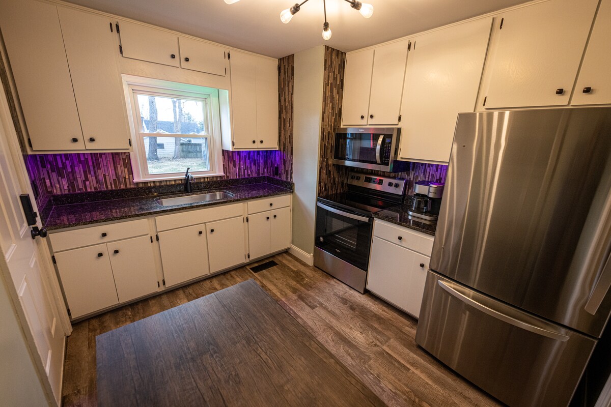 The kitchen features modern appliances, including a stainless steel refrigerator and oven. Ample counter space is complemented by bright cabinets and a striking backsplash with purple accent lighting. A window allows natural light to fill the room and offers a view of the outdoor area.