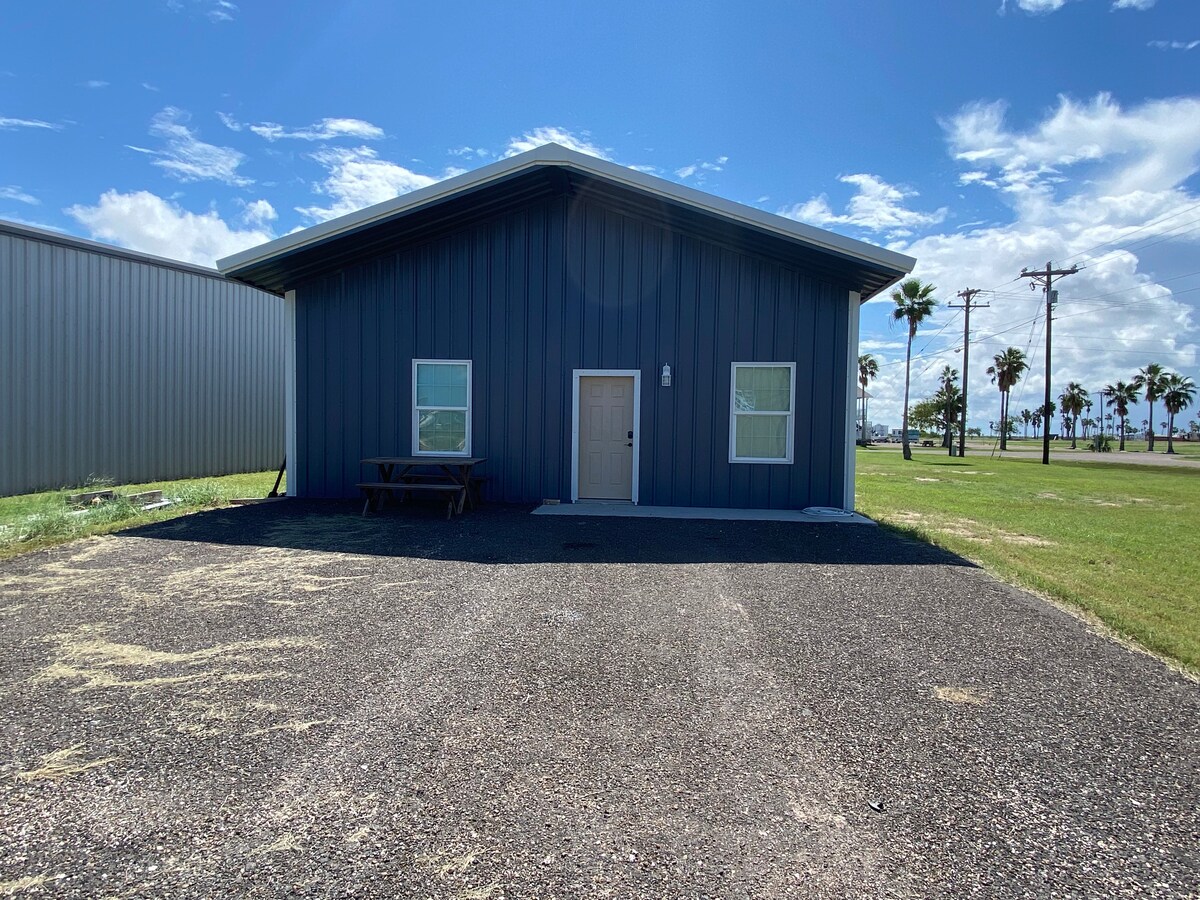 The exterior of the efficiency apartment is presented, showcasing a modern blue structure with a simple entrance. Flanking trees and cloudy skies can be seen, while a gravel driveway leads up to the building. Nearby, wide open spaces contribute to a serene environment.