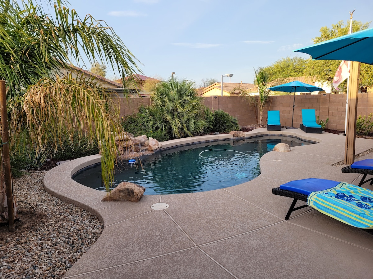 The pool area is surrounded by lush greenery and features a curved pool shape with natural stone accents. Two blue lounge chairs are positioned on the side, along with a colorful patterned mat. A vibrant blue umbrella provides shade over the seating area.