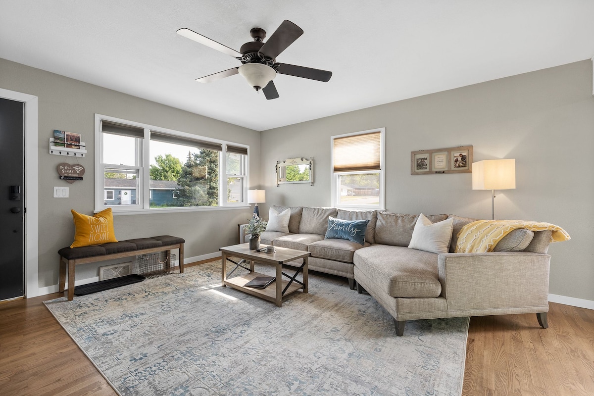 A modern living area features a spacious sectional sofa, complemented by a decorative throw pillow and a light-colored area rug. Natural light filters through multiple windows, and a ceiling fan adds to the comfort of the space. A coffee table and a bench are also visible.
