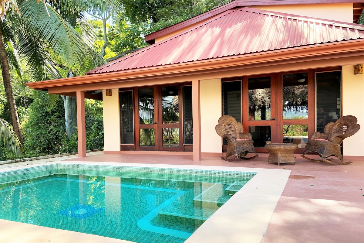 A private pool is framed by a spacious patio, with a villa featuring large glass windows and a red metal roof. Two wicker chairs are positioned near the pool, providing a relaxing spot to enjoy the surrounding greenery.
