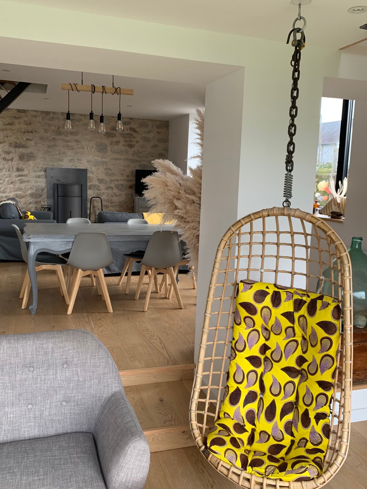A stylish hanging chair features a vibrant yellow and brown patterned blanket draped over it, adding a touch of color. The background reveals a dining area with a wooden table and modern chairs, accompanied by light-colored stone walls, creating a contemporary yet warm environment.