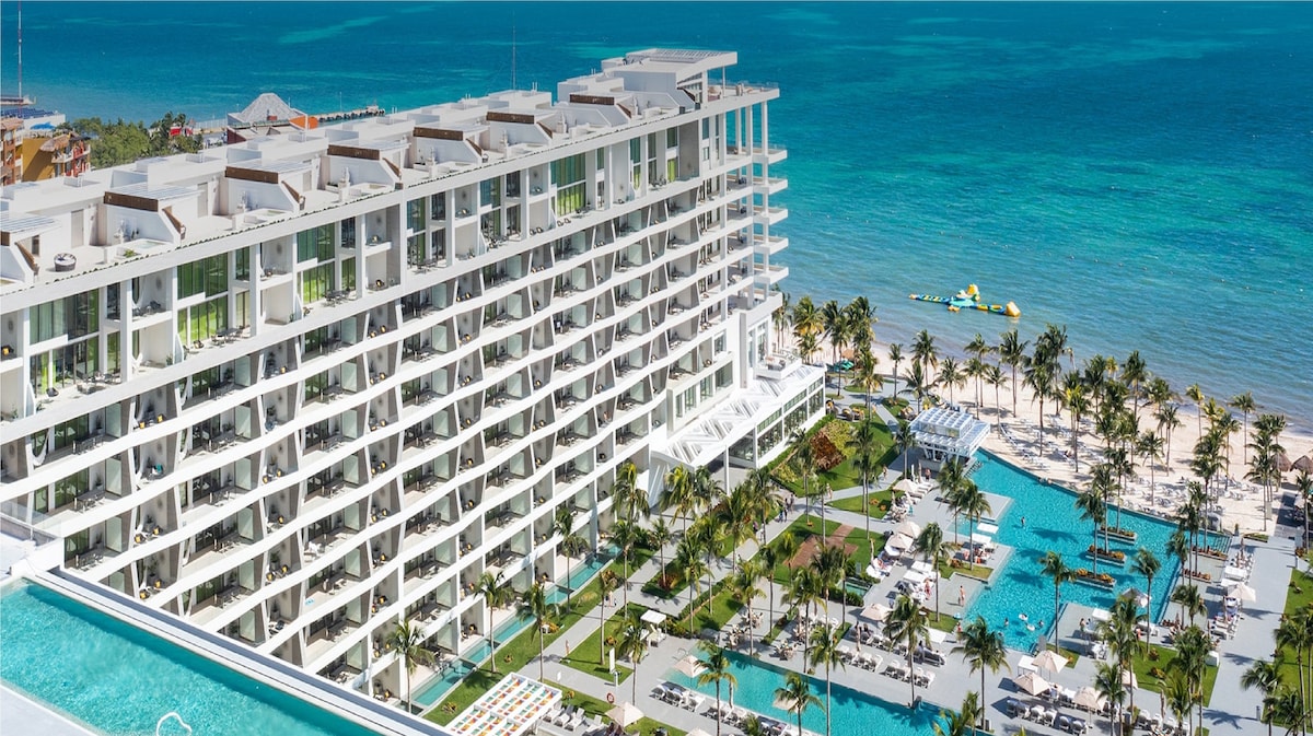 An aerial view showcases the contemporary architecture of the resort, featuring multiple floors with expansive balconies. The vibrant blue waters of the Caribbean Sea contrast with the lush greenery surrounding the expansive pool area and white sandy beach, highlighting a luxurious vacation atmosphere.