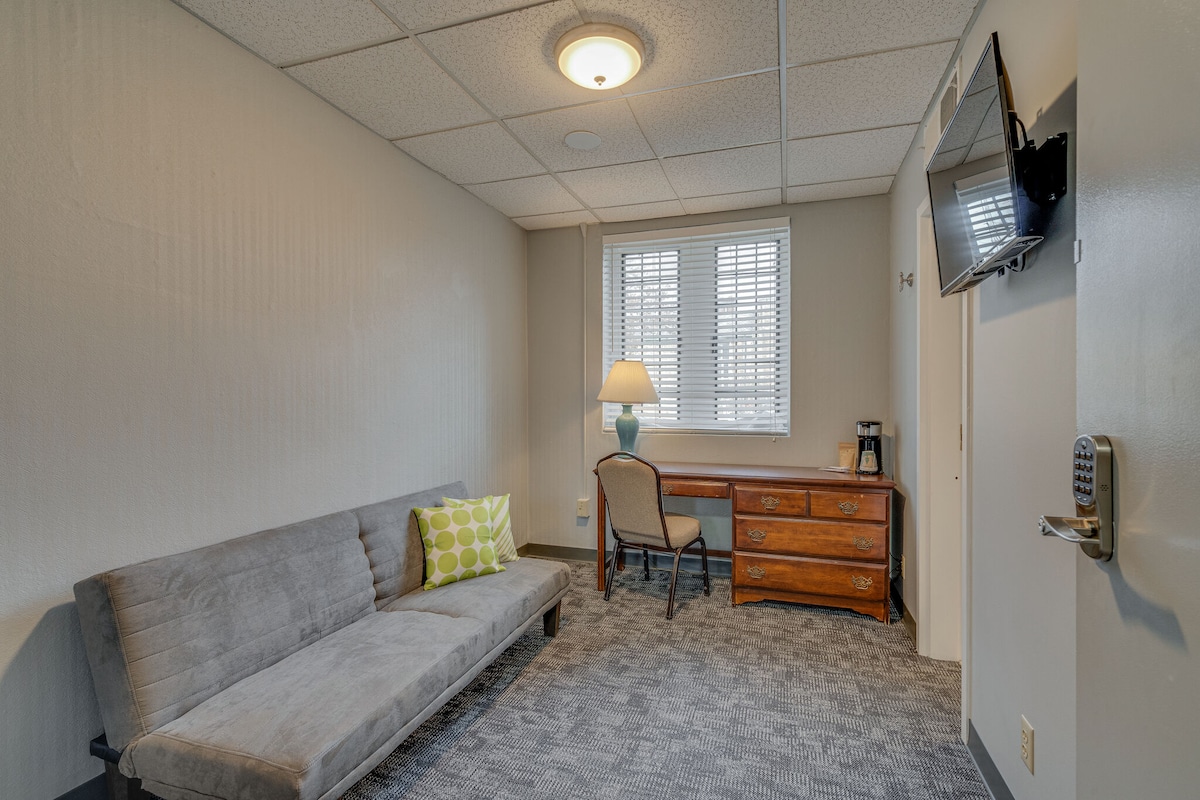 A living space is shown, featuring a gray futon, a wooden desk with a chair, and a lamp. The wall is adorned with a window with blinds, allowing natural light, while a television is mounted on the opposite wall.