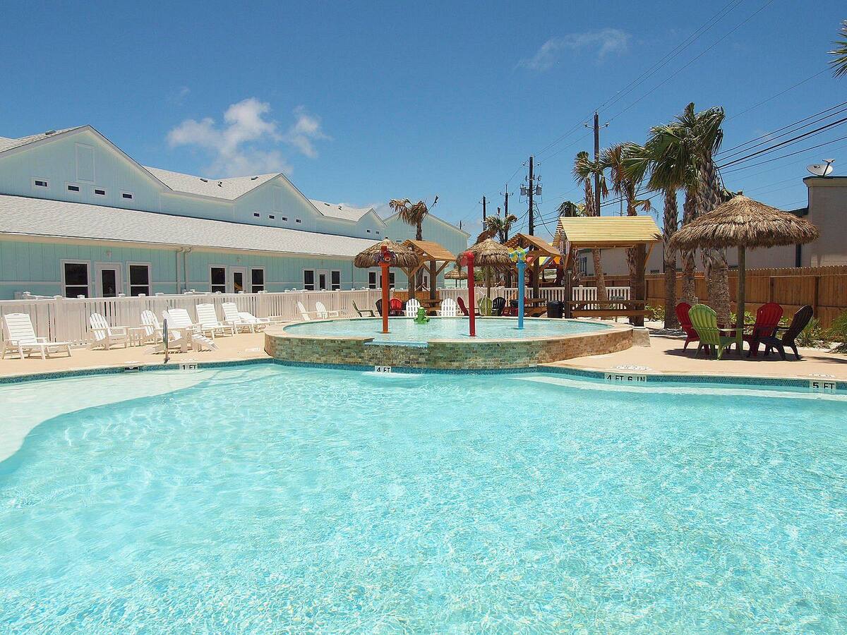 A resort pool area is highlighted, featuring a large swimming pool with clear blue water. Surrounding the pool are colorful umbrellas and lounge chairs. Palm trees are visible in the background, offering a tropical feel. A shallow area within the pool is equipped for children.