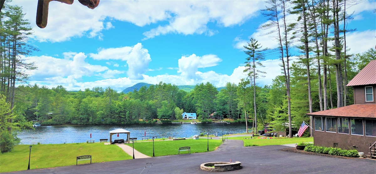 A serene lakeside view is presented, featuring a calm body of water surrounded by lush greenery and tall trees. A gentle pathway leads towards the water, with a fire pit situated on a spacious lawn that extends toward the water's edge.
