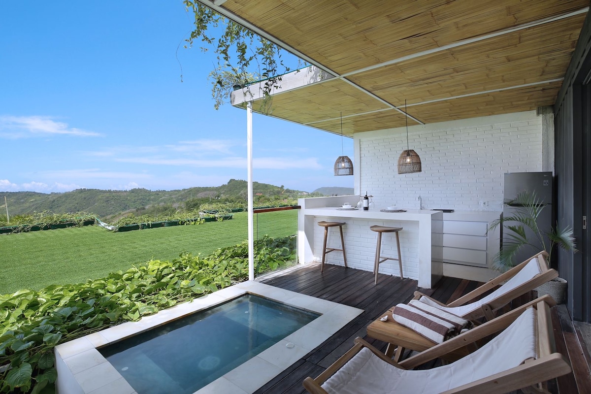 A private outdoor space features a small jacuzzi surrounded by wooden decking. Two lounge chairs are positioned nearby, with a modern kitchen bar visible under an overhanging bamboo roof. Lush greenery and rolling hills are seen in the background against a bright blue sky.