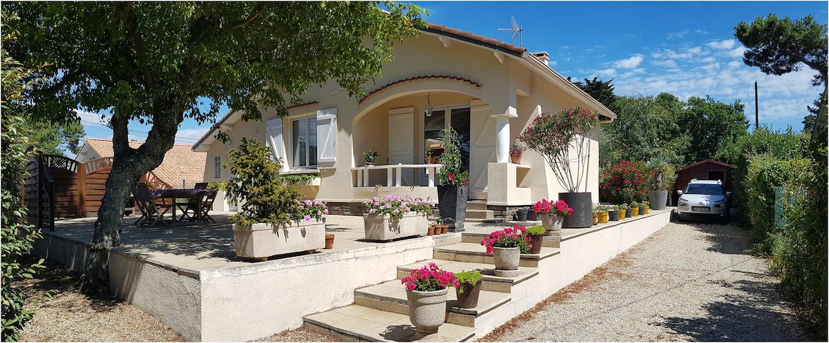 A classic-style villa is surrounded by greenery, featuring stone steps leading to a welcoming entrance. Flower pots adorn the steps, adding color. The structure includes large windows and a covered porch area for outdoor relaxation.